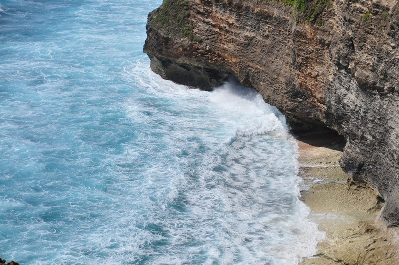 Turquoise ocean waves crashing against layered sedimentary cliff face with green vegetation on top